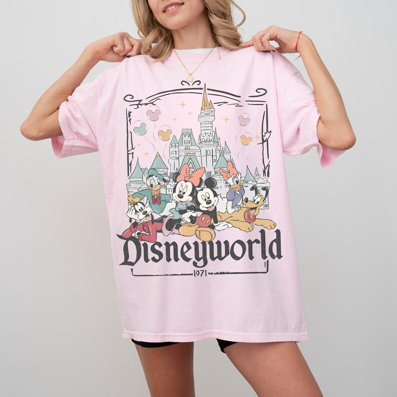 May include: A light pink t-shirt featuring a graphic of Disney characters including Mickey and Minnie Mouse, Donald Duck, and Goofy. The word "Disneyworld" is printed in a stylized font, with "1971" below.