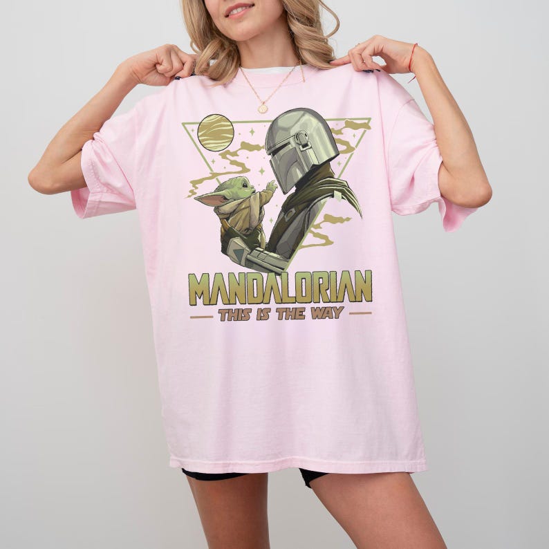 May include: A light pink t-shirt featuring a graphic of The Mandalorian and Grogu. The design includes a helmet, a baby Grogu, and the text "MANDALORIAN - THIS IS THE WAY" in green and gold.