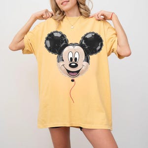 Puede incluir: Camiseta amarilla con un dise&ntilde;o de globo de Mickey Mouse. El globo es negro, blanco y beige con una cuerda roja. La camiseta es de color amarillo liso.