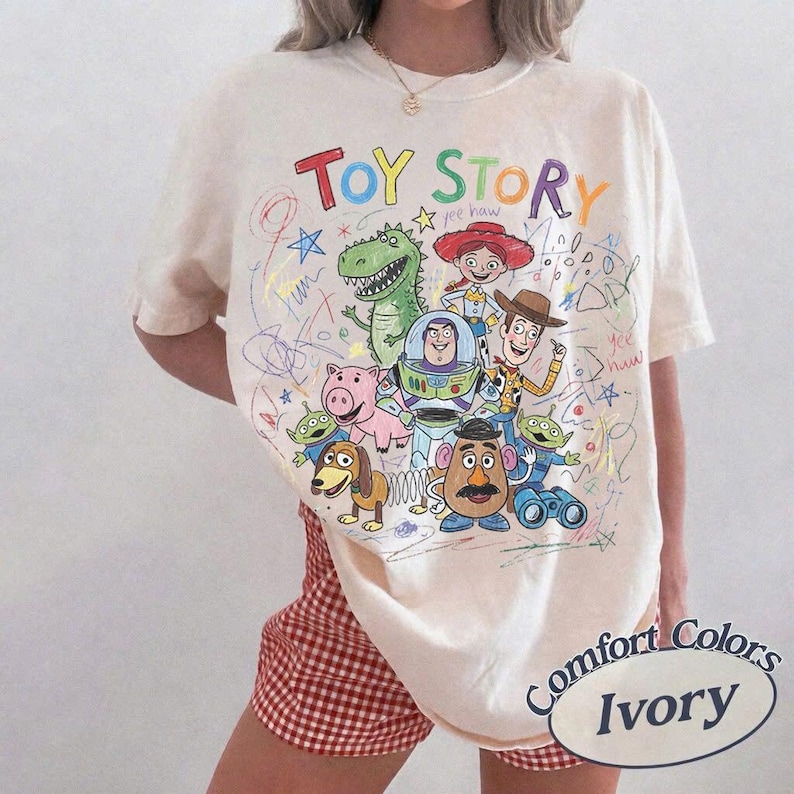 May include: Ivory-colored t-shirt featuring a colorful "Toy Story" graphic with various characters and the text "Toy Story" in rainbow colors. The shirt is a Comfort Colors brand.