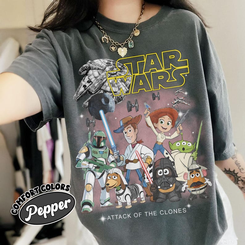 May include: Dark grey t-shirt with a Star Wars graphic, featuring characters from the film and Toy Story. The design includes the text "STAR WARS" and "ATTACK OF THE CLONES". The shirt has a Comfort Colors Pepper label.