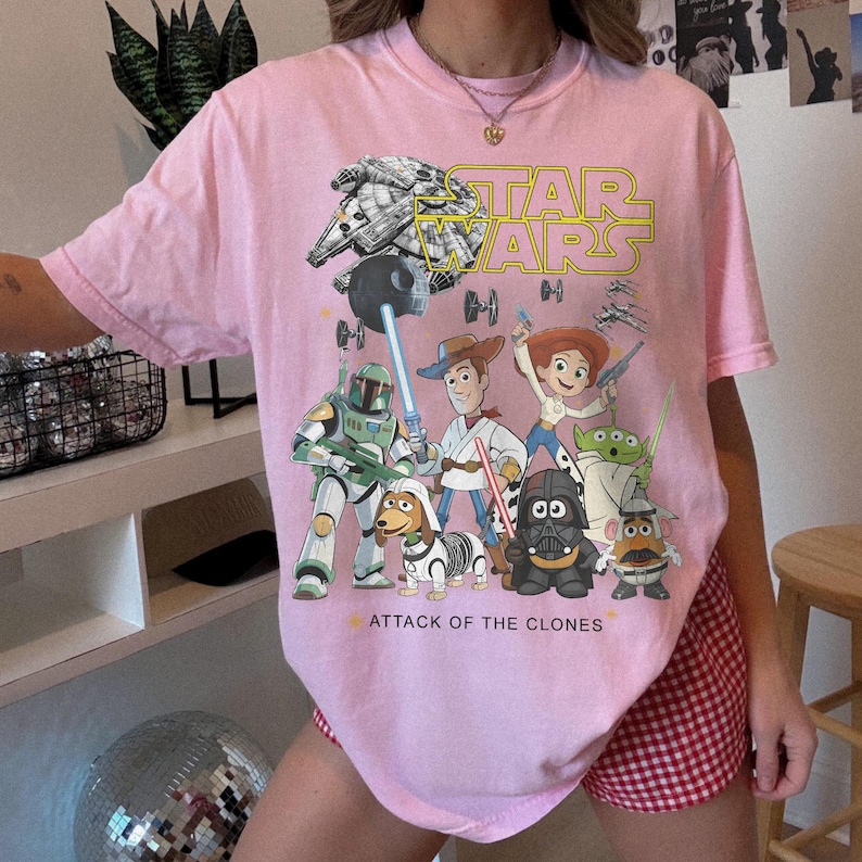 May include: Pale pink t-shirt with a Star Wars graphic. The design includes the Millennium Falcon, Darth Vader, and other characters, with the words "STAR WARS" in yellow. The text "ATTACK OF THE CLONES" is also visible on the shirt.