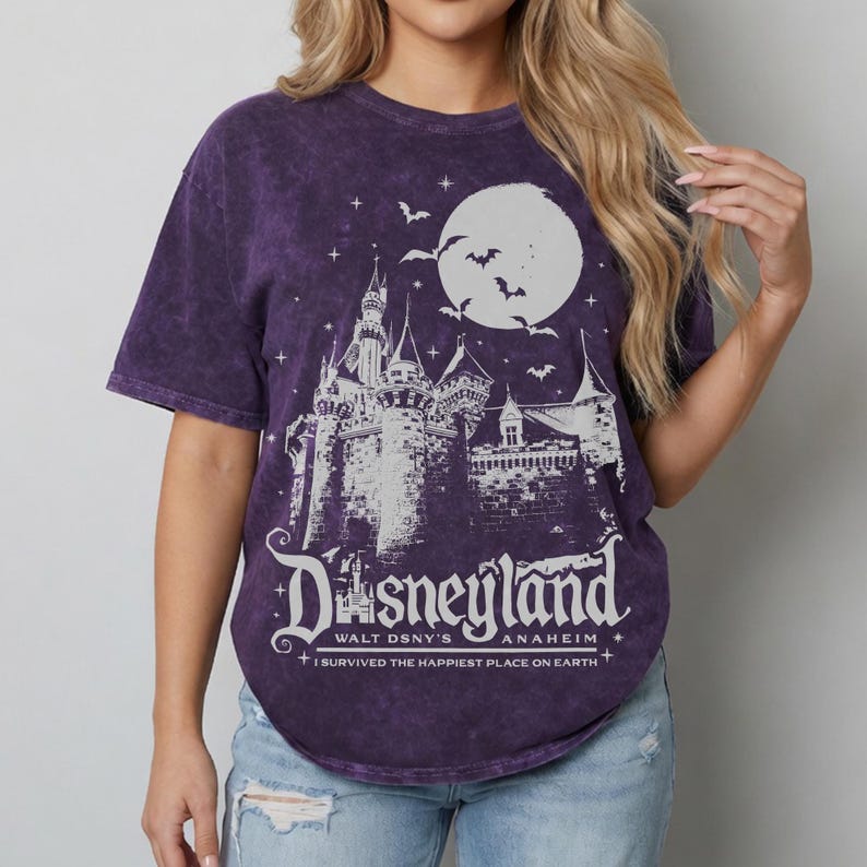 Pode incluir: Camiseta roxa com um gr&aacute;fico branco de um castelo, lua e morcegos. O texto "Disneyland Walt DSNY'S Anaheim" est&aacute; impresso abaixo do castelo, com a frase "I survived the happiest place on Earth".