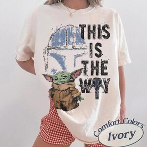 May include: Ivory-colored t-shirt featuring a graphic of a green alien character, a blue helmet, and the text "THIS IS THE WAY." The design includes a brown jacket and a black helmet. The shirt is from Comfort Colors.
