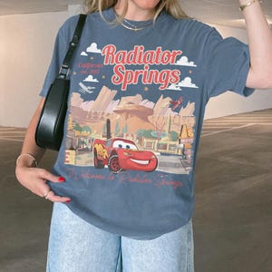 May include: A blue t-shirt featuring a graphic of the town of Radiator Springs from the movie Cars. The design includes the text "Radiator Springs" and "Welcome to Radiator Springs", along with an image of Lightning McQueen.