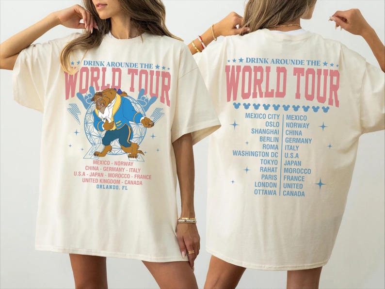 K&ouml;nnte beinhalten: Cremefarbenes T-Shirt mit dem Text "DRINK AROUND THE WORLD TOUR" in Rosa. Auf der Vorderseite befindet sich eine Cartoonfigur in einem blauen Mantel. Auf der R&uuml;ckseite sind St&auml;dte wie Mexiko-Stadt, Oslo und Shanghai aufgef&uuml;hrt.