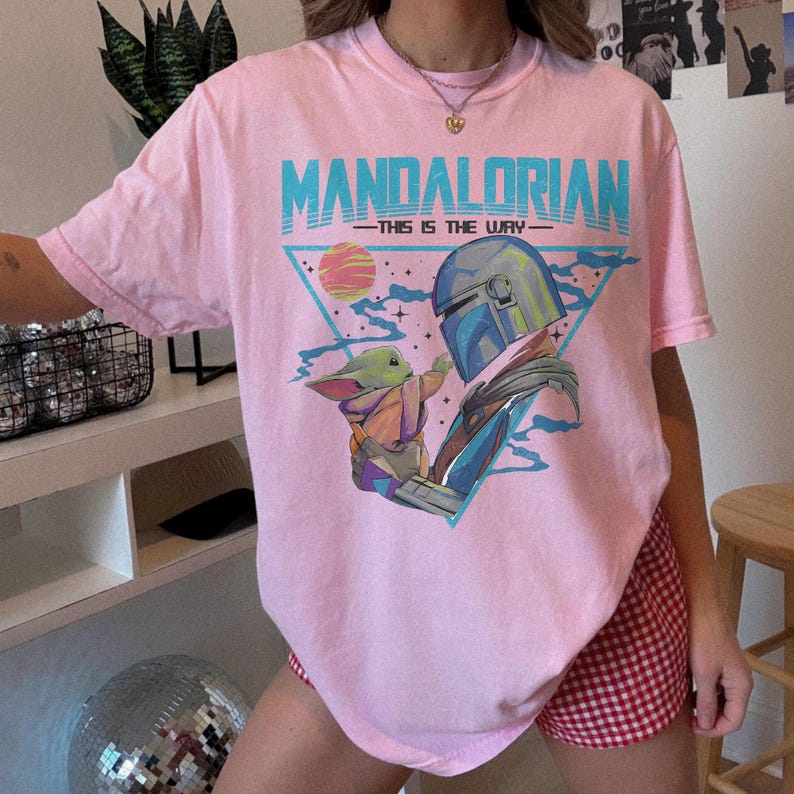 May include: Pink t-shirt featuring a graphic of The Mandalorian and Grogu, with the text "MANDALORIAN - THIS IS THE WAY" in blue. The design includes a triangle, a planet, and a space scene. The shirt is a short-sleeved, crew neck style.
