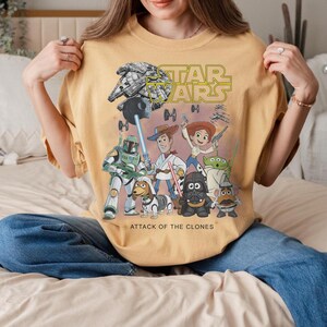 May include: Mustard yellow t-shirt with a mashup design of Star Wars and Toy Story characters. The design includes the Star Wars logo, a Millennium Falcon, and characters like Woody, Buzz Lightyear, and Darth Vader. The text "ATTACK OF THE CLONES" is printed below the characters.