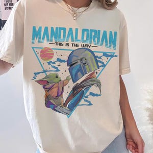 May include: Cream-colored t-shirt featuring a graphic of The Mandalorian and Grogu, with the text "MANDALORIAN - THIS IS THE WAY" in blue. The design includes a helmeted figure and a small green creature, set against a triangle and a planet.