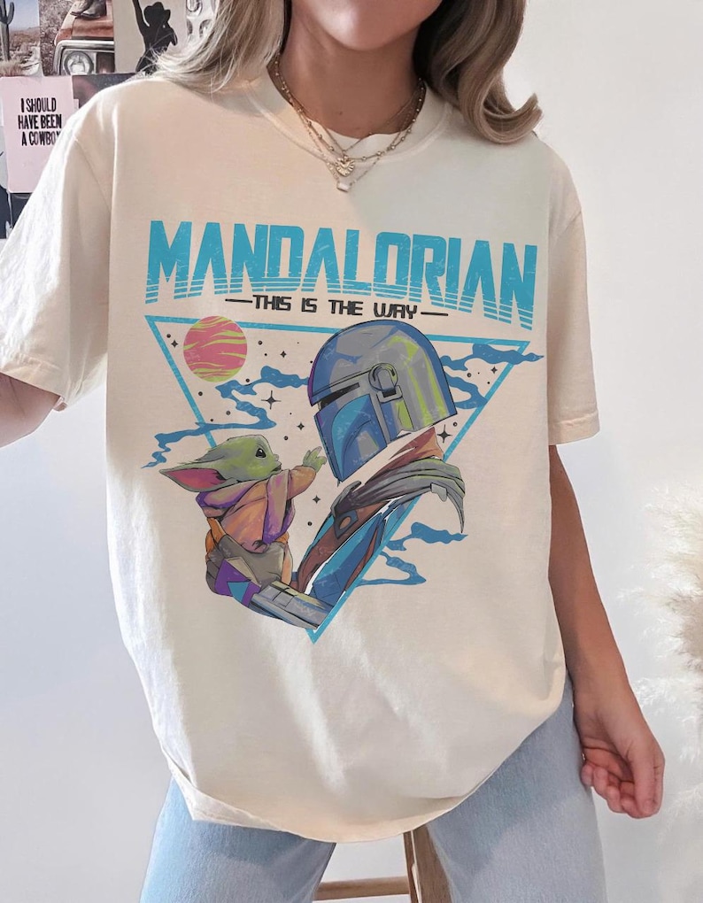 May include: Cream-colored t-shirt featuring a graphic of The Mandalorian and Grogu, with the text "MANDALORIAN - THIS IS THE WAY" in blue. The design includes a helmeted figure and a small green creature, set against a triangle and a planet.