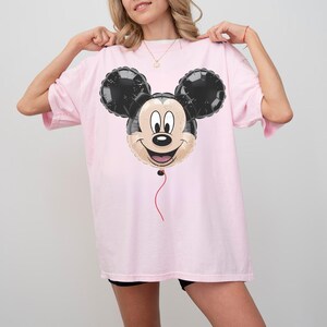 Puede incluir: Camiseta holgada rosa claro con un dise&ntilde;o de globo de Mickey Mouse. El globo tiene orejas negras y una cara sonriente. Una cuerda roja cuelga de la parte inferior. La camiseta es de estilo casual.