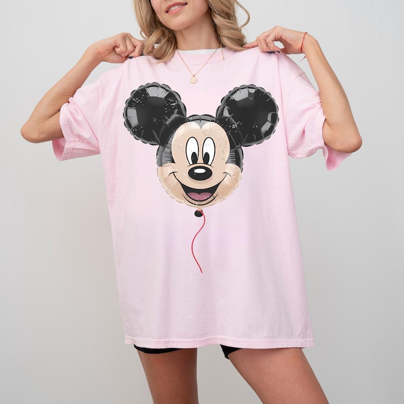 Puede incluir: Camiseta holgada rosa claro con un dise&ntilde;o de globo de Mickey Mouse. El globo tiene orejas negras y una cara sonriente. Una cuerda roja cuelga de la parte inferior. La camiseta es de estilo casual.