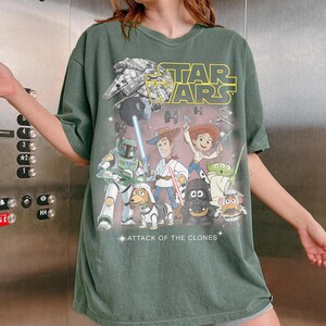 May include: Green t-shirt with a Star Wars graphic, including the text "STAR WARS" in yellow. The design features characters from Star Wars and Toy Story. The words "ATTACK OF THE CLONES" are also visible.