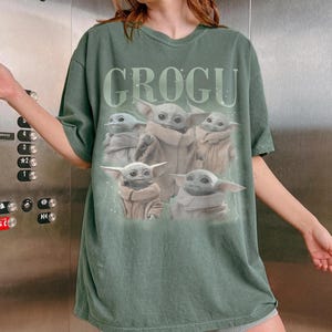 May include: Green t-shirt featuring the word "GROGU" above a graphic of Grogu, also known as Baby Yoda, from the Star Wars series. The graphic shows Grogu in various poses. The shirt has short sleeves and a relaxed fit.