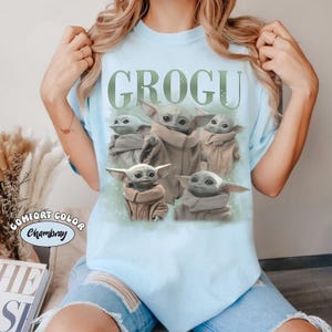 Puede incluir: Camiseta azul claro con la palabra "GROGU" en verde sobre un gr&aacute;fico de Grogu, tambi&eacute;n conocido como Baby Yoda, en varias poses. La camiseta tiene una etiqueta "Comfort Color Chambray".