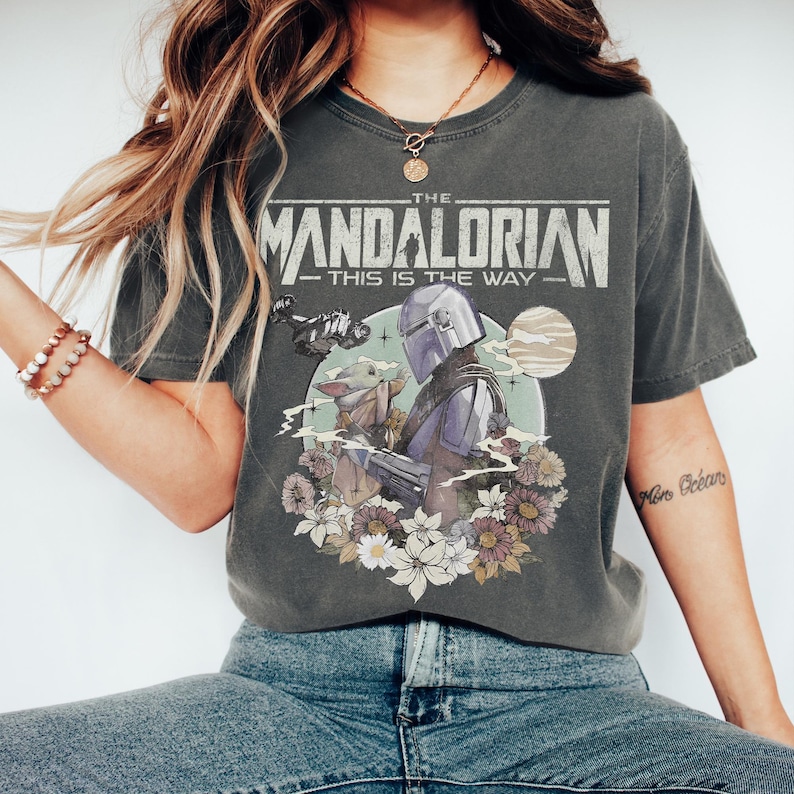 May include: Dark gray t-shirt featuring a graphic from The Mandalorian. The design includes the text "THE MANDALORIAN - THIS IS THE WAY" above an illustration of the Mandalorian, Grogu, and flowers. The shirt is a vintage style.