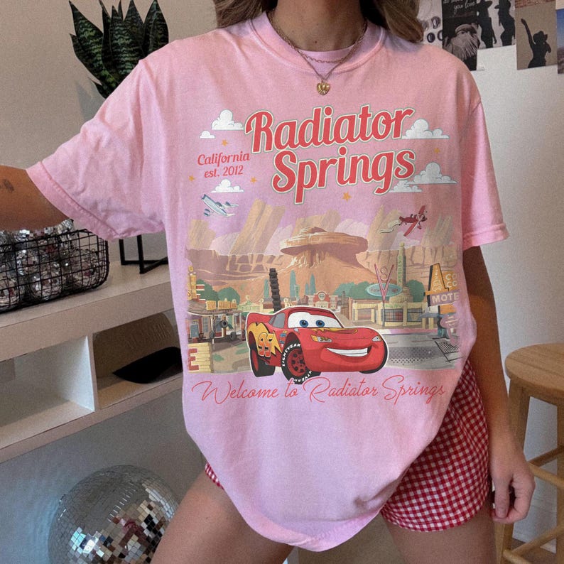 May include: Pink t-shirt featuring a graphic of Radiator Springs from the movie Cars. The design includes the text "Radiator Springs" and "Welcome to Radiator Springs". The shirt also has the text "California est. 2012".