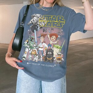 May include: Blue t-shirt with a Star Wars and Toy Story mashup graphic. The design includes the text "STAR WARS" and "ATTACK OF THE CLONES", featuring various characters from both franchises.