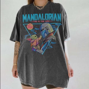 May include: Dark gray t-shirt featuring a graphic of The Mandalorian and Grogu, with the text "MANDALORIAN - THIS IS THE WAY" in blue. The design includes a triangle with a planet and stars.