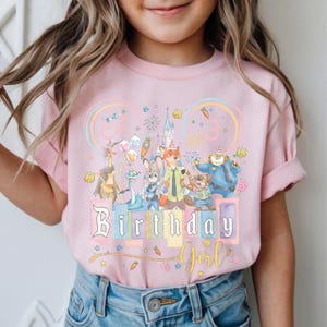 May include: Pale pink t-shirt with a colourful graphic of cartoon characters, a castle, and the words "Birthday Girl." The design includes a rainbow, stars, and the number "3."