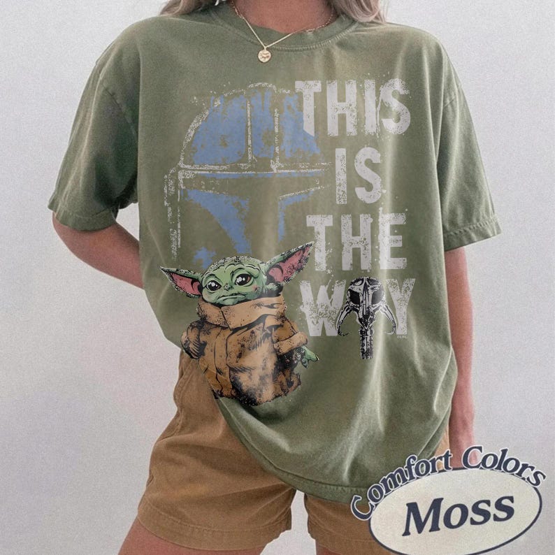 May include: Olive green t-shirt featuring a graphic of Grogu, a Mandalorian helmet, and the phrase "THIS IS THE WAY" in white. The shirt is a Comfort Colors brand in the color Moss.