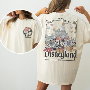 May include: Cream-colored t-shirt featuring a Minnie Mouse graphic on the front and a Disneyland design on the back. The Disneyland graphic includes Mickey Mouse, Donald Duck, and other characters, with the text "Disneyland 1955".