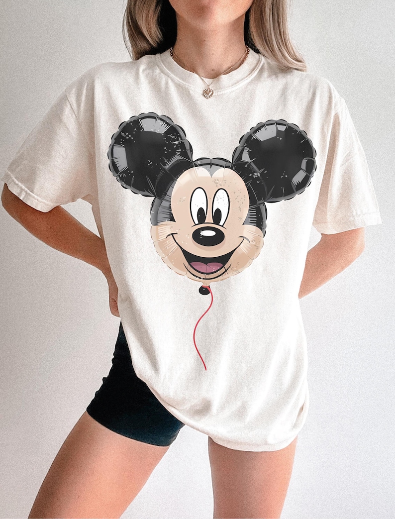 Puede incluir: Camiseta color crema con un dise&ntilde;o de globo de Mickey Mouse. El globo tiene orejas negras y una cara sonriente con una cuerda roja. La camiseta es de corte holgado.
