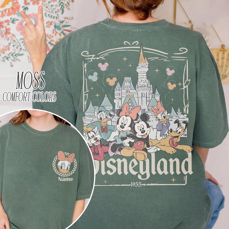 May include: Moss green t-shirt with a Disneyland graphic on the back, featuring Mickey and Minnie Mouse, Donald Duck, and Goofy. The front of the shirt has a small Daisy Duck graphic. The words "Comfort Colors" are printed on the left side.