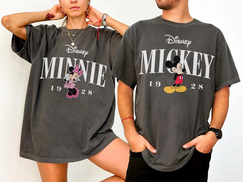 May include: Two charcoal grey t-shirts featuring Disney characters. One shirt displays "MINNIE" with Minnie Mouse in a pink dress, and the other shows "MICKEY" with Mickey Mouse. Both shirts include the year "1928".