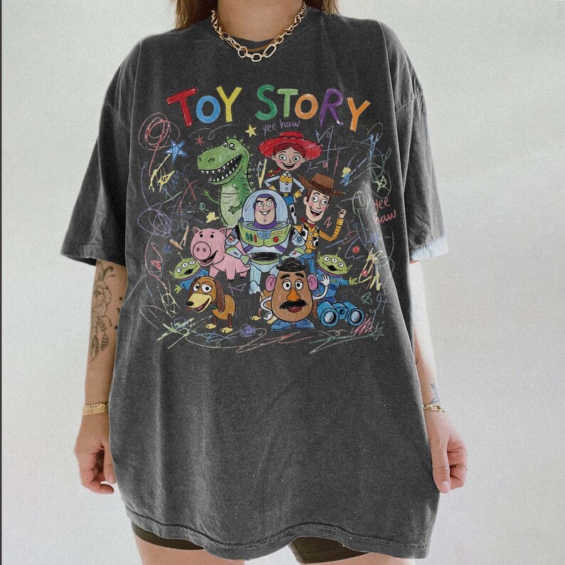 May include: Dark gray t-shirt featuring the words "TOY STORY" in colorful letters above a graphic of characters from the animated film. The design includes Woody, Buzz Lightyear, Rex, and other characters. The shirt has a relaxed fit.