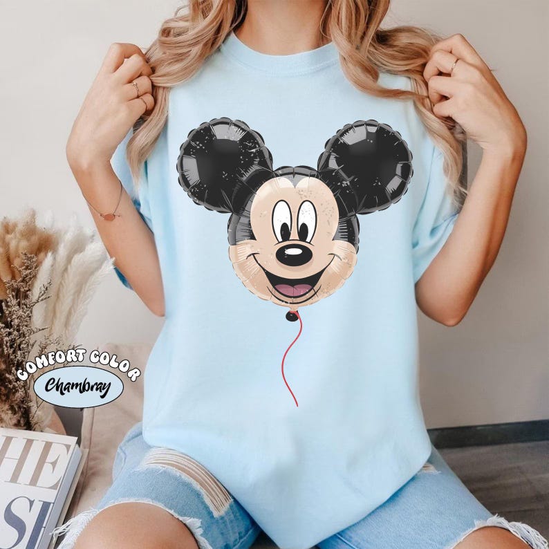 Puede incluir: Camiseta azul claro con un dise&ntilde;o de globo de Mickey Mouse. El globo tiene orejas negras y una cara sonriente. La camiseta tiene la inscripci&oacute;n "COMFORT COLOR Chambray".
