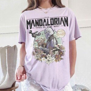 May include: A light purple t-shirt featuring a graphic of The Mandalorian and Grogu surrounded by flowers, with the text "THE MANDALORIAN - THIS IS THE WAY" in black.