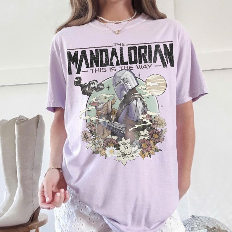 May include: A light purple t-shirt featuring a graphic of The Mandalorian and Grogu surrounded by flowers, with the text "THE MANDALORIAN - THIS IS THE WAY" in black.