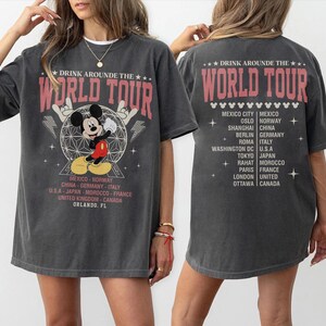 K&ouml;nnte beinhalten: Dunkelgraues T-Shirt mit einem Mickey Mouse-Motiv und dem Text "DRINK AROUND THE WORLD TOUR". Das Shirt listet St&auml;dte wie Berlin, Tokio und Rom auf. Auf der R&uuml;ckseite des Shirts steht ebenfalls "DRINK AROUND THE WORLD TOUR" und eine Liste von St&auml;dten.