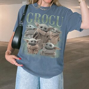 Puede incluir: Camiseta azul con la palabra "GROGU" en verde sobre un gr&aacute;fico de Grogu, tambi&eacute;n conocido como Baby Yoda, de The Mandalorian. El gr&aacute;fico muestra m&uacute;ltiples im&aacute;genes de Grogu con una capa marr&oacute;n.
