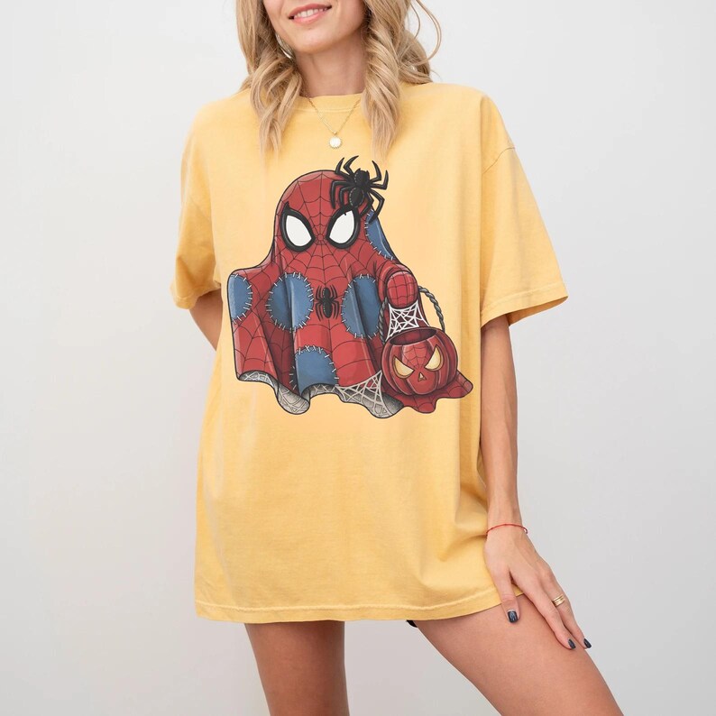 May include: Yellow t-shirt featuring a Halloween-themed graphic. The graphic depicts a ghost in a Spider-Man costume holding a pumpkin-shaped candy bucket. A black spider sits atop the ghost's head. The shirt is a casual, oversized fit.