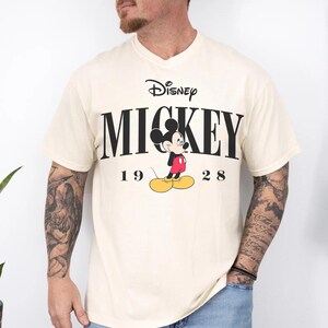 May include: A cream-coloured t-shirt with the Disney logo and the word "MICKEY" in bold black letters. The image includes a graphic of Mickey Mouse in his classic outfit, with the year "1928" below the name. The shirt has a V-neck.