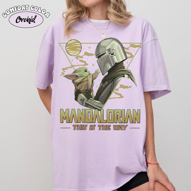 May include: A light purple t-shirt featuring a graphic of The Mandalorian and Grogu. The design includes the text "MANDALORIAN" and "THIS IS THE WAY" in gold lettering. The shirt has the "Comfort Colors" and "Orchid" logo.