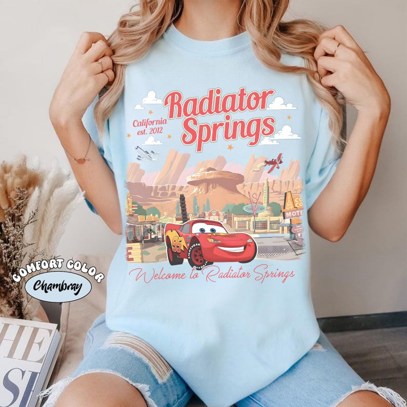 May include: Light blue t-shirt with a graphic of Radiator Springs, featuring a red race car and desert landscape. Text includes "Radiator Springs," "California est. 2012," and "Welcome to Radiator Springs."