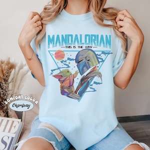 May include: Light blue t-shirt featuring a graphic of The Mandalorian and Grogu, with the text "MANDALORIAN" and "THIS IS THE WAY" in blue. The design includes a triangle, a sunset, and stars. The shirt is labeled "Comfort Color Chambray".