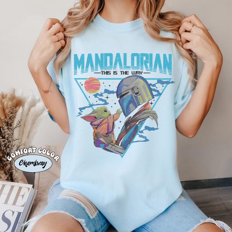 May include: Light blue t-shirt featuring a graphic of The Mandalorian and Grogu, with the text "MANDALORIAN" and "THIS IS THE WAY" in blue. The design includes a triangle, a sunset, and stars. The shirt is labeled "Comfort Color Chambray".