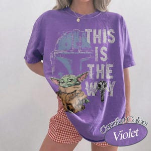 May include: Violet Comfort Colors t-shirt featuring a graphic of the Mandalorian helmet, Baby Yoda, and the phrase "This is the Way." The design includes a helmet and a mask. The shirt is a short-sleeved crew neck.