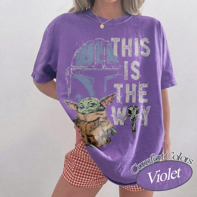 May include: Violet Comfort Colors t-shirt featuring a graphic of the Mandalorian helmet, Baby Yoda, and the phrase "This is the Way." The design includes a helmet and a mask. The shirt is a short-sleeved crew neck.