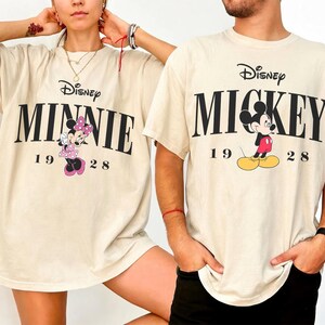 May include: Two cream-coloured t-shirts featuring Disney characters. One shirt displays "MINNIE" with a cartoon Minnie Mouse, and the other says "MICKEY" with Mickey Mouse. Both shirts include the year "1928" and the Disney logo.