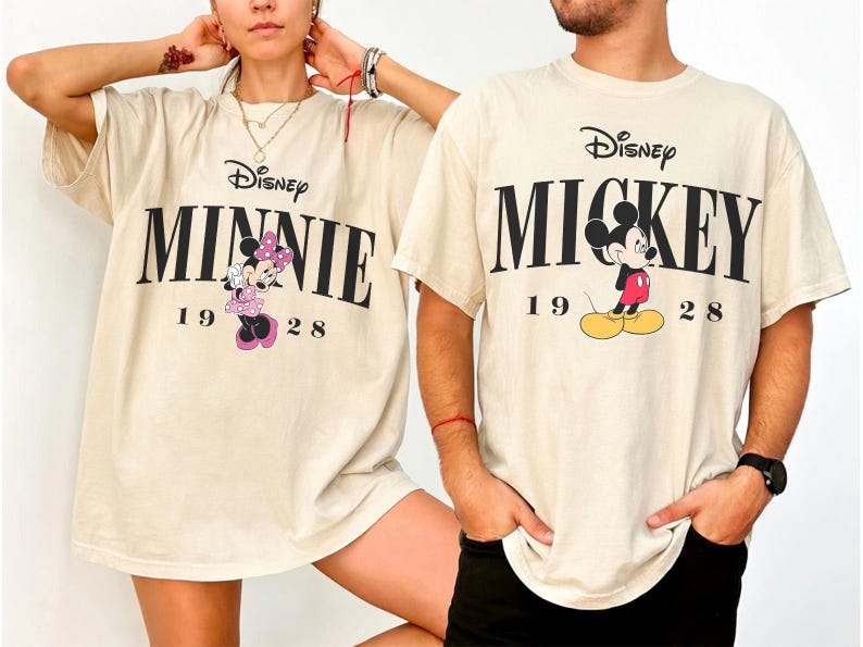 May include: Two cream-coloured t-shirts featuring Disney characters. One shirt displays "MINNIE" with a cartoon Minnie Mouse, and the other says "MICKEY" with Mickey Mouse. Both shirts include the year "1928" and the Disney logo.