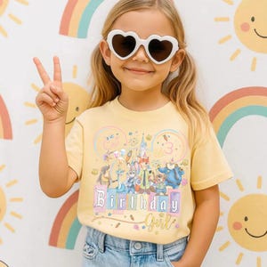 May include: A young girl wearing heart-shaped sunglasses and a yellow t-shirt with a colourful graphic. The shirt features the text "Birthday Girl" and a cartoon illustration. The background includes a rainbow and suns.