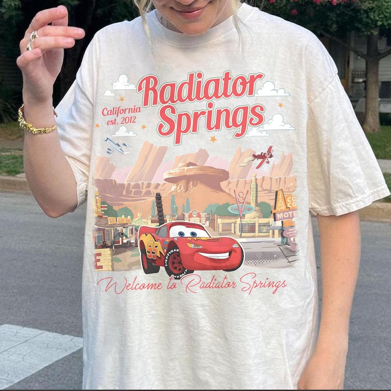 May include: Cream-colored t-shirt featuring a graphic of Radiator Springs with Lightning McQueen. The design includes the text "Radiator Springs" in red, "California est. 2012", and "Welcome to Radiator Springs".