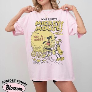 May include: A light pink t-shirt featuring a vintage cartoon graphic of Mickey Mouse in "Get a Horse!". The design includes Mickey, a horse, and a cart in yellow and black. The shirt also has the words "Comfort Colors" and "Blossom".