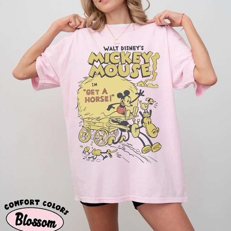 May include: A light pink t-shirt featuring a vintage cartoon graphic of Mickey Mouse in "Get a Horse!". The design includes Mickey, a horse, and a cart in yellow and black. The shirt also has the words "Comfort Colors" and "Blossom".