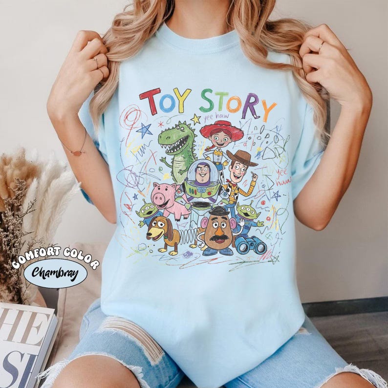 May include: Light blue t-shirt featuring the words "TOY STORY" in colorful letters, with a graphic of characters from the movie. The design includes Woody, Buzz Lightyear, and other characters. The shirt is a Comfort Color Chambray.